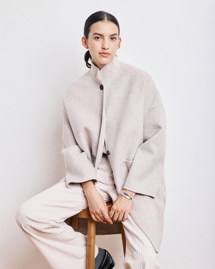 Person wearing a light gray coat sitting on a stool against a white background