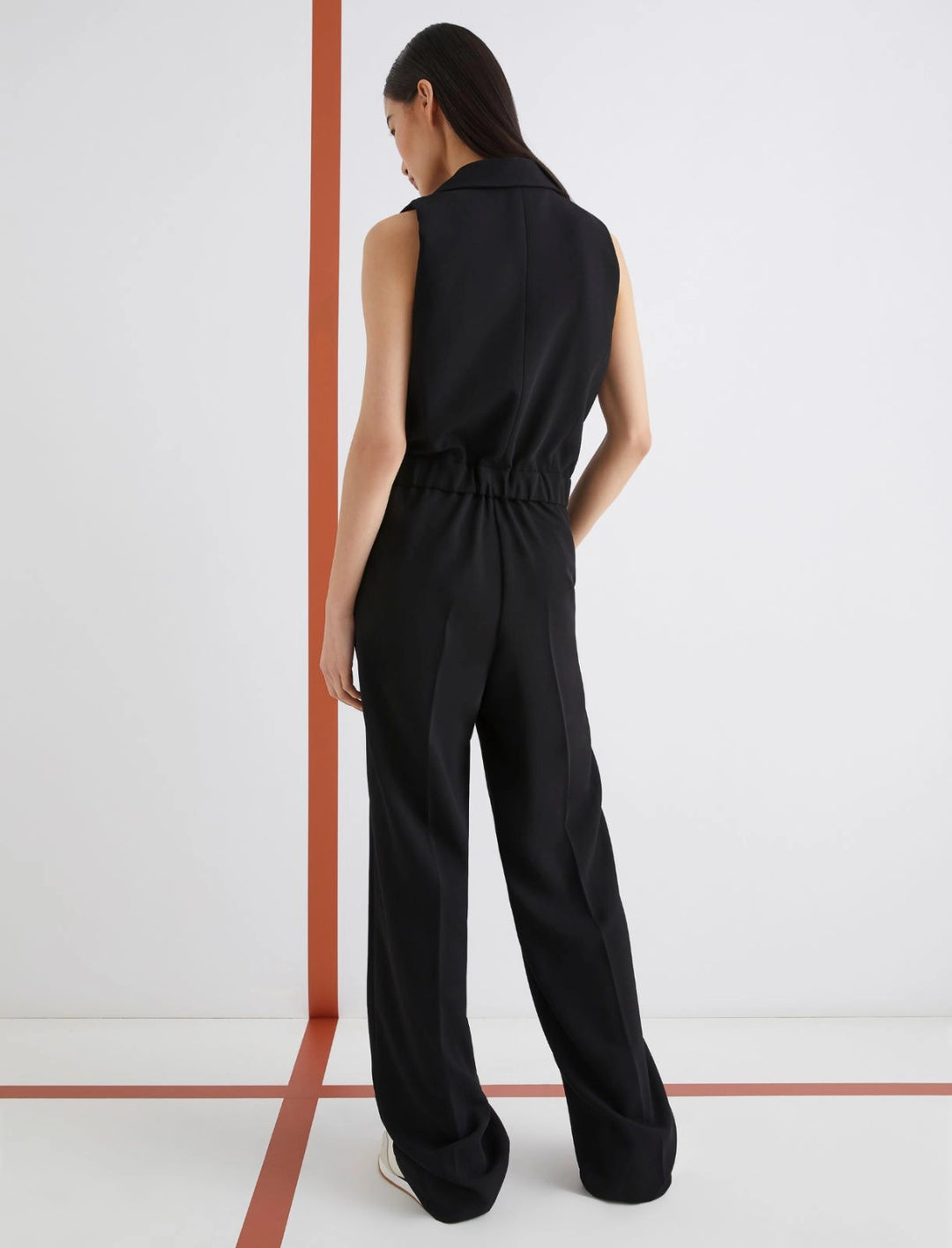 Woman wearing a black jumpsuit standing against a white wall.