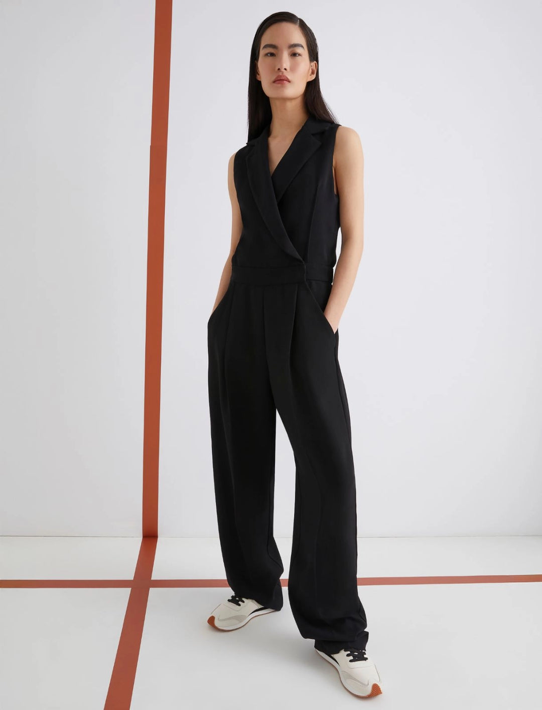 Woman wearing a black jumpsuit standing in a minimalistic room with a white wall and red floor.