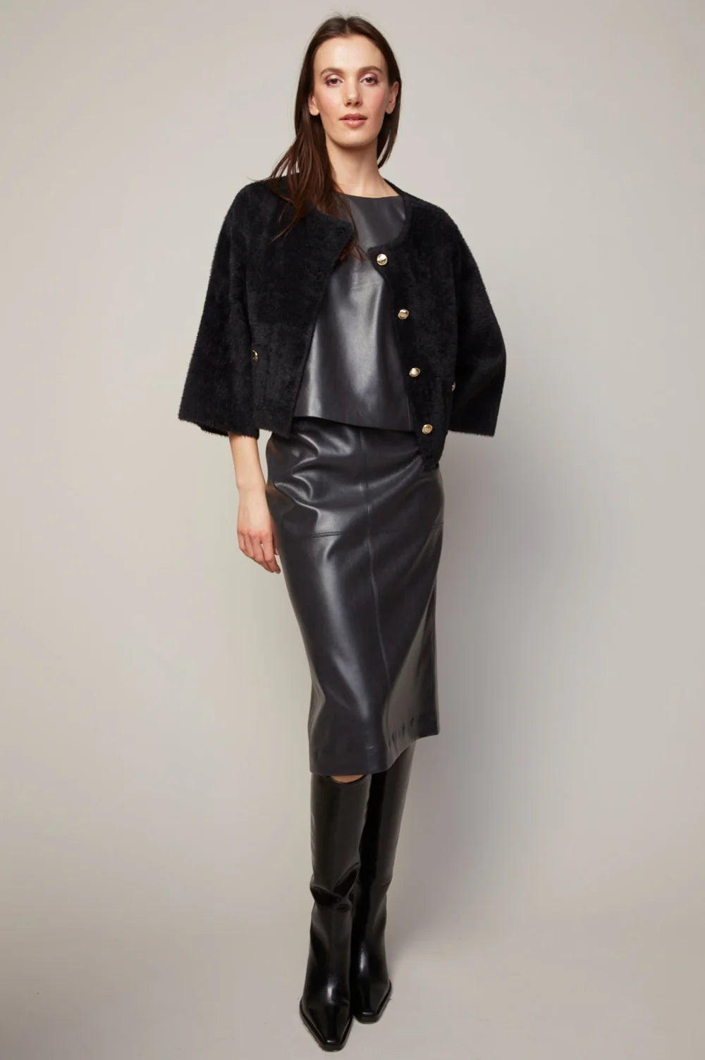Woman wearing a black leather outfit with a jacket, skirt, and boots on a plain background