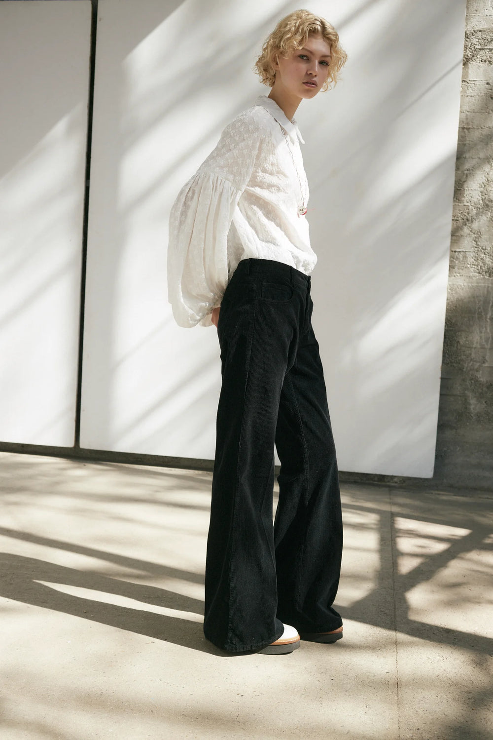 Person wearing a white blouse and black pants standing against a light-colored wall.