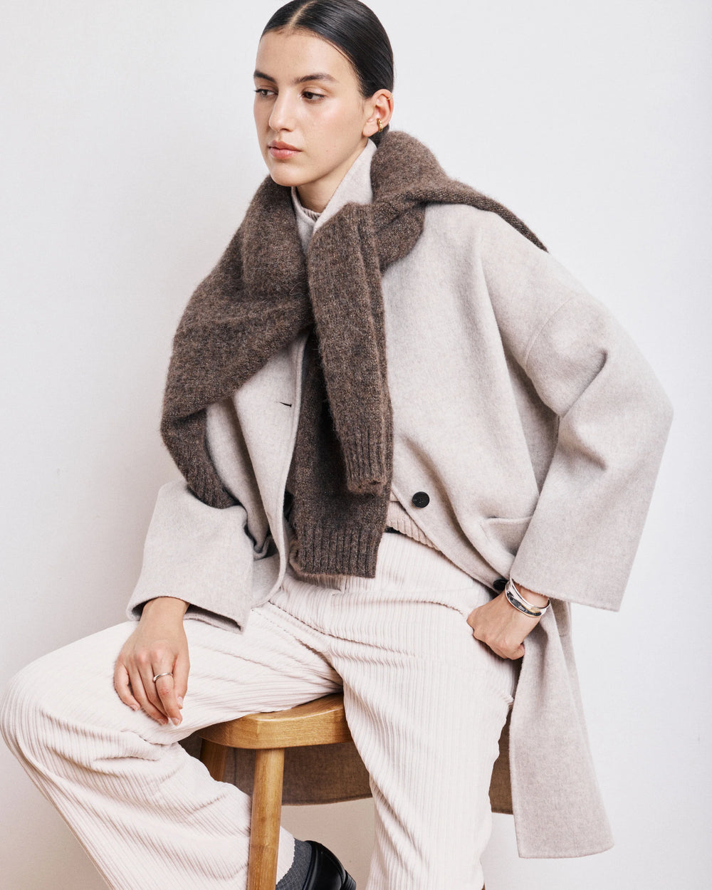 Person wearing a light gray coat and white pants with a brown scarf, sitting on a wooden stool against a white background.
