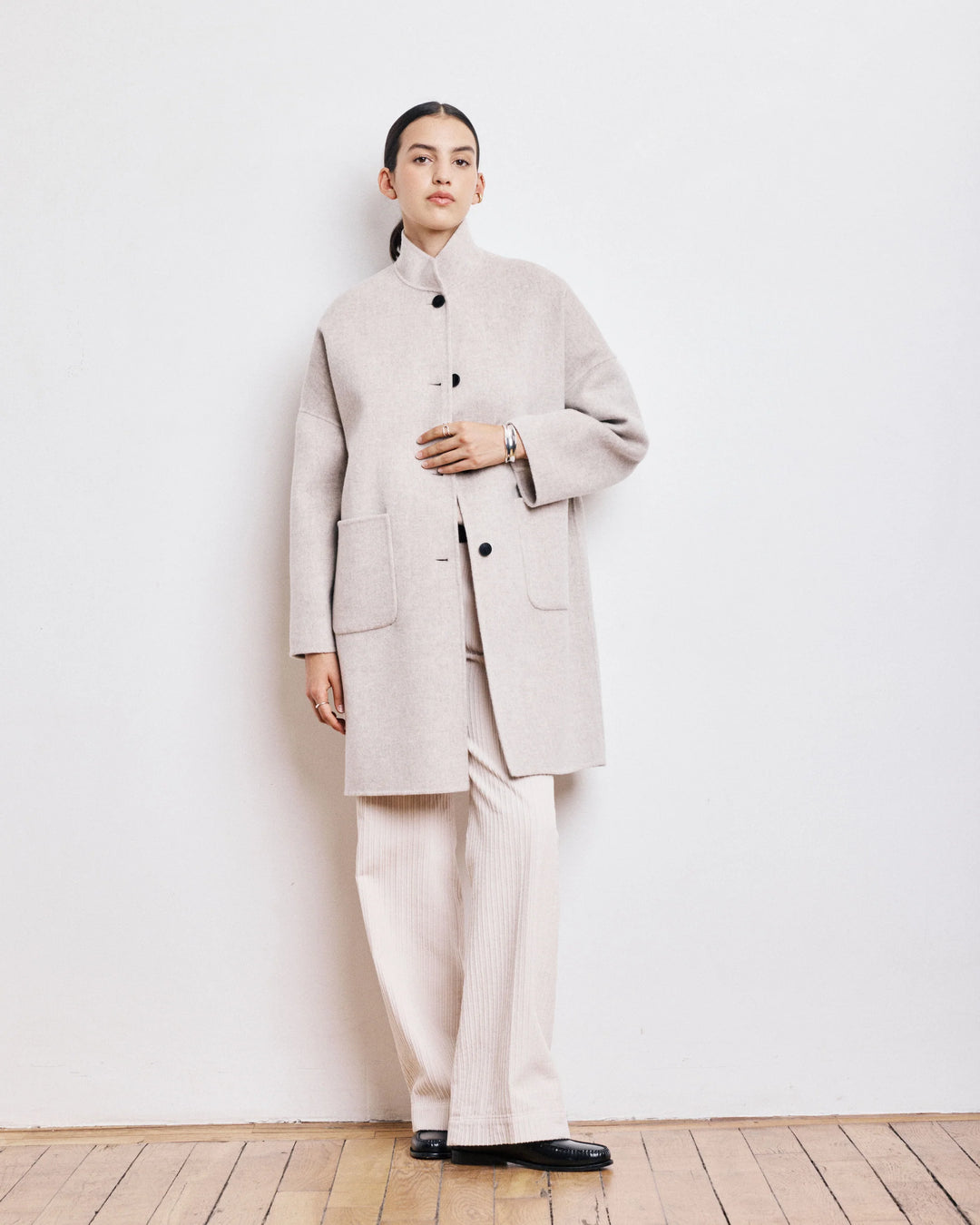 Person wearing a beige coat and pants standing against a white wall.